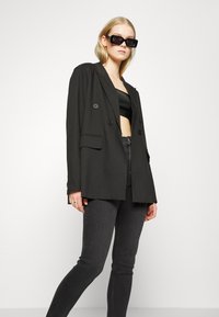 Blazer preto com design de duplo botão, colarinho em lapela e tecido texturizado, combinado com um top preto cropped e jeans escuros.