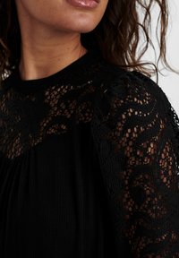 Top nero caratterizzato da un tessuto testurizzato con dettagli in pizzo sulle spalle, che presenta motivi floreali e intricate aperture. Design a maniche corte.