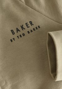 Khaki kangast, millel on sile tekstuur, sisaldab tumedat logo: "BAKER BY TED BAKER." Disain sisaldab lihtsaid jooni ja tihedat istuvust.