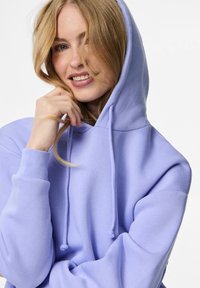 Ung kvinne som smiler, iført en lys lilla hettejakke med hetten opp, og som berører håret sitt med høyre hånd mot en hvit bakgrunn.