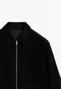 Schwarze Wildlederjacke mit kurzem Kragen, silbernem Reißverschluss vorne und flachen Nähten. Glatte Textur, minimales Design.