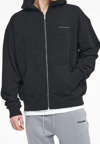 Schwarze Zip-Hoodie mit geripptem Saum und Bündchen, Fronttaschen und Logo-Druck auf der Brust. Weicher Stoff mit lockerer Passform und Kapuze.
