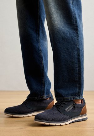 Vyrų sportiniai bateliai su tamsiai mėlynu tinkleliu, rudos odos akcentais, guminėmis padais ir struktūrizuotu dizainu. Suderinti su tamsiai mėlynomis džinsais.