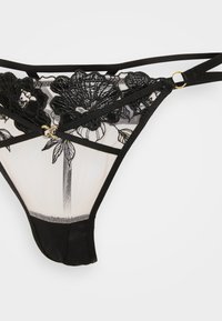 Schwarzes Spitzen- und Netz-String mit floralem Stickerei, dünnen Trägern sowie Details aus goldfarbenem Ring und Doppelring auf weißem Hintergrund.
