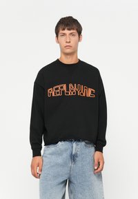 Felpa nera con il testo arancione "REPLAYING". Presenta un collo a giro e una vestibilità comoda. Abbinata a jeans in denim azzurro chiaro.