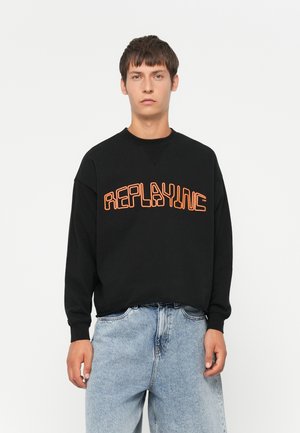 Black sweatshirt with orange "REPLAYING" text. Features a crew neckline and relaxed fit. Paired with light blue denim jeans.