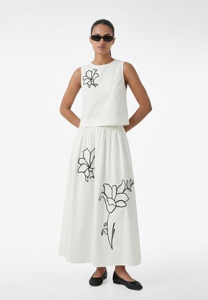 Donna che indossa una canottiera bianca e una gonna lunga con contorni floreali neri, scarpe basse nere e occhiali da sole rettangolari neri, in piedi con le mani dietro la schiena.