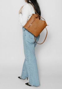 Zaino in finta pelle marrone con texture, caratterizzato da una chiusura con zip, mostrato su uno sfondo chiaro, indossato sulla schiena con una lunga tracolla.