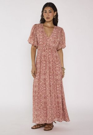 Femme debout sur fond blanc, portant une robe longue rose à motifs floraux avec des manches courtes, des sandales brunes et des bijoux dorés.