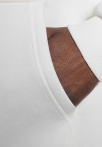Un tissu de pull blanc avec un col arrondi et des poignets côtelés. Une main brune apparaît, tendue vers une poche latérale, montrant un contraste de texture.