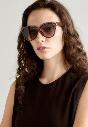 Kilpkonnakujulised prillid suurte, nurgeliste raamide ja tumedate läätsedega. Kandjal on pikad, lokkis juuksed ja seljas on ilma varrukateta must topp.