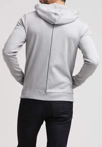Sudadera con capucha gris con un detalle de costura en la parte posterior, hecha de una tela suave. Presenta mangas largas y un corte regular, combinada con jeans oscuros.