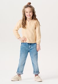 Jasnokremowa bluzka z długim rękawem i teksturowanym dekoltem w kształcie litery V, zestawiona z jasnoniebieskimi dżinsami z mankietami i beżowymi trampkami. Dziecko stoi z rękami na biodrach.