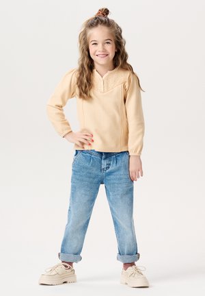 Helles beige Langarmshirt mit strukturiertem V-Ausschnitt, kombiniert mit hellblauen, umgeschlagenen Jeans und beigen Turnschuhen. Das Kind steht mit auf die Hüften gestützten Händen.