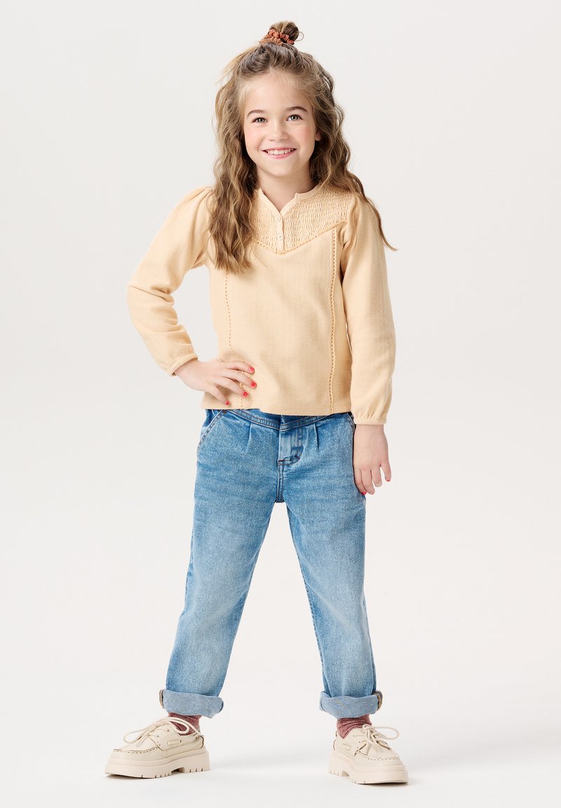 Jasnokremowa bluzka z długim rękawem i teksturowanym dekoltem w kształcie litery V, zestawiona z jasnoniebieskimi dżinsami z mankietami i beżowymi trampkami. Dziecko stoi z rękami na biodrach.