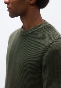 Zeleni pleteni pulover z rebrasto teksturo, okrogel izrez in oprijet kroj. Prikaz iz profila poudarja brezšivni detajl na ramenu.