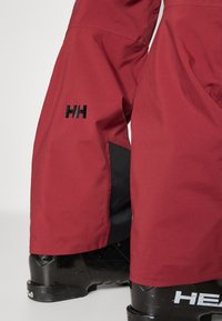 Pantalons imperméables rouges avec des accents noirs et un logo sur la jambe. Présentent une texture lisse et un design ajusté, adaptés aux activités d'hiver.