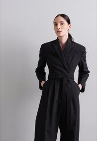 Salopetă neagră, ajustată, cu un design înfășurat, având talie cu curea, mâneci lungi și buzunare laterale, fabricată dintr-un material texturat.