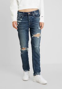 Jean en denim bleu déchiré avec une coupe slim, présentant des détails vieillis au niveau des genoux et un léger motif de délavage. Associés à des baskets blanches.