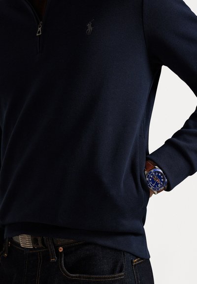 Marineblå genser med halvdels glidelås og liten logo-detalle, parret med en blå klokke med metallring og brun lærrem.