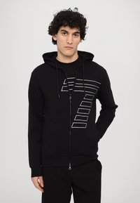 Čierna mikina na zips vyrobená z mäkkej tkaniny, so sivou grafickou potlačou na prednej strane, s kapucňou s dresovým podšívkou a prednými vreckami.
