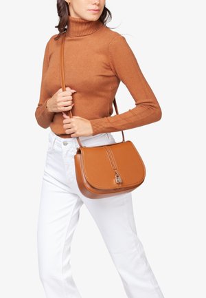 Sac à bandoulière en cuir marron au design structuré, avec un rabat avant, des détails en zip et un accent de verrouillage en argent. Porté avec un pantalon blanc.