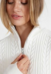 Fehér cable-knit pulóver magas gallérral és ezüst cipzárral. A mandzsetták bordázottak, és a szöveten kiemelkedő kötött textúra látható.