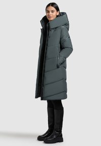 Long puffer coat in dark gray, with a high collar and chevron stitching design. Features zippered pockets and a slim fit silhouette.