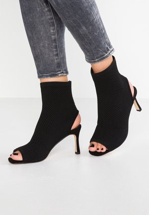 Fødder iført sorte teksturerede ankelstøvler med åbne tæer og åbne hæle, kombineret med grå, op rullede skinny jeans.