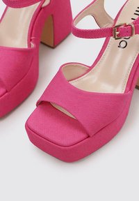 Sandalias de tacón alto rosas con parte superior de tela, que presentan un diseño de cintas cruzadas, puntera cuadrada y un tacón grueso. Acabado texturizado cerca de las puntas.