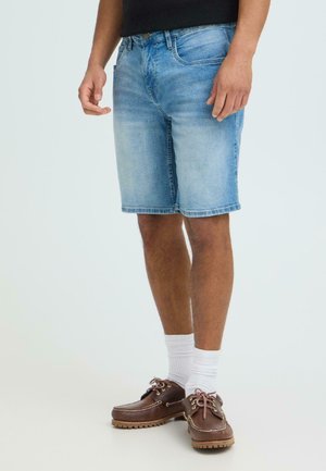 Mann iført lyseblå denimshorts, hvite sokker, brune skinnbåtsko og svart skjorte, står på hvit bakgrunn.