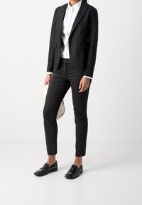 Blazer negro a medida sobre una camisa blanca, combinado con pantalones negros de corte ajustado y mocasines negros. Lleva un bolso de color claro.
