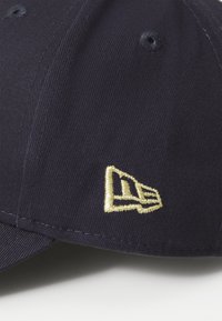Navy baseball cap with a structured crown, featuring a gold embroidered logo on the side and textured fabric. Smooth visor.
