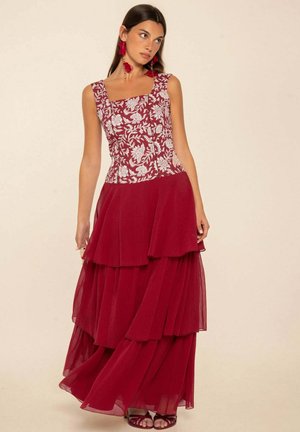Mujer con un vestido rojo de falda escalonada y cuerpo blanco bordado con flores, de pie sobre un fondo beige.