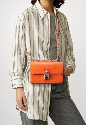 Bolso de cuero naranja con acabado texturizado, con cierre de cerradura plateada y correa de cadena, usado con una camisa de rayas de gran tamaño.