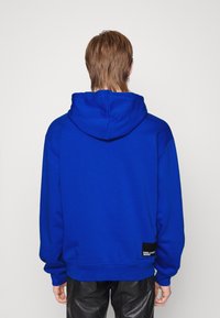 Sudadera de color azul real con capucha ajustable, puños y dobladillo acanalados. Presenta una pequeña etiqueta de logo negra en la parte inferior izquierda. Tejido suave.