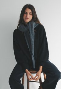 Overdimensioneret sort sweater med tekstureret stof, parret med et kontrasterende gråt tørklæde. Model sidder på en træskammel med hænderne hvilende på knæene.