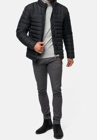 Schwarze Daunenjacke mit Reißverschluss und Stehkragen; grauer Pullover darunter; graue Slim-Fit-Hose; schwarze Knöchelstiefel.