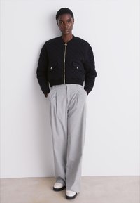 Casaco bomber preto acolchoado com fecho dourado e bolsos frontais, combinado com calças pregueadas cinza claro e sapatos preto e branco.