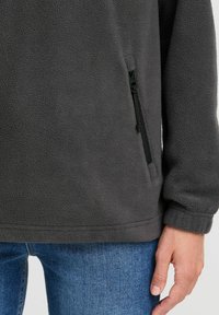 Pullover grigio scuro in pile con una texture morbida, dotato di tasca laterale con zip e polsini aderenti, abbinato a jeans blu.