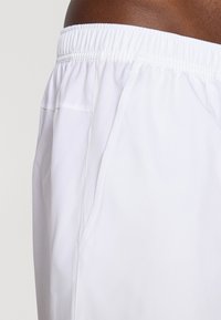Shorts de sport blancs avec une taille élastique, dotés d'une poche latérale et d'une texture lisse, conçus pour le confort et la liberté de mouvement.
