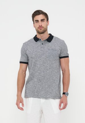 Mężczyzna w krótkim, szarym polo z czarnym kołnierzykiem i mankietami, białych spodniach i czarnym zegarku na nadgarstku, stojący na białym tle.