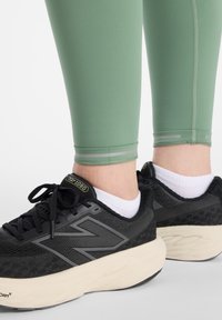 Leggings deportivos verdes con detalles reflectantes, combinados con zapatillas de correr negras con detalles grises y una suela acolchada color crema.