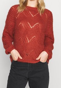 Chunky roter Strickpullover mit offenem Zickzackmuster, Rundhalsausschnitt, übergroßen Ärmeln und geripptem Saum. Kombiniert mit schwarzen Jeans.