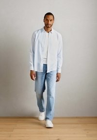 Mann, der auf einem Holzfußboden vor einer schlichten Wand geht, gekleidet in ein hellblau gestreiftes Hemd, ein weißes T-Shirt, hellblaue Jeans und weiße Sneaker.