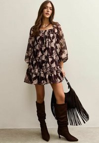 Vestido de gasa floral en púrpura oscuro con mangas abullonadas, dobladillo con volantes, combinado con botas marrones hasta la rodilla y un bolso negro con flecos.