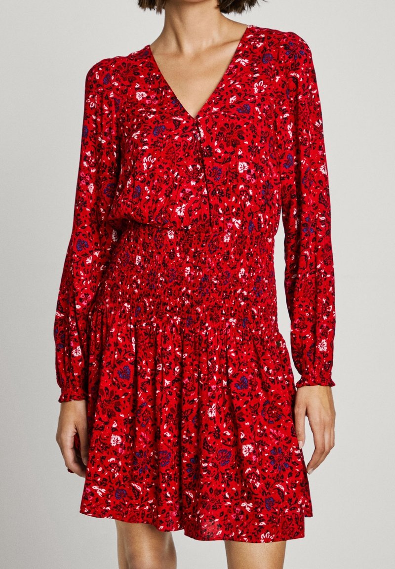 Robe rouge à manches longues avec motif floral, col en V, taille froncée et jupe évasée, portée par une personne debout sur un fond uni.