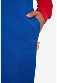 Main reposant dans la poche d’un vêtement bleu avec une étiquette orange visible de la marque Opposuits, portant une chemise rouge à manches longues.