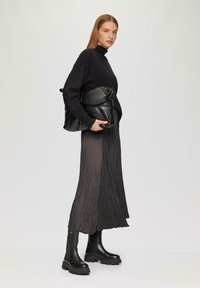 Un pull noir à col montant, une jupe plissée noire et des bottes noires robustes. La mannequin tient un sac noir texturé. Arrière-plan neutre.