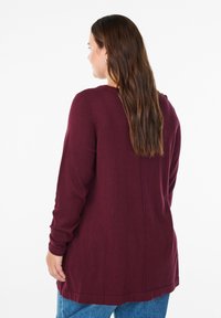 Burgunderfarbener Langarm-Cardigan aus weichem Strickmaterial, mit geradem Saum und einfachem Design, über einer blauen Jeans getragen.